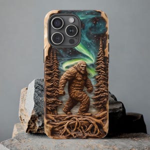 May include: A wooden phone case featuring a carved Bigfoot design. The case has a detailed relief of the creature walking through a forest with a starry sky and aurora borealis in the background. The case is brown and has a natural wood grain.