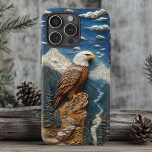 Puede incluir: Una funda para teléfono con un diseño tallado en 3D de un águila calva posada en la cima de una montaña. El fondo presenta un cielo azul con nubes blancas y una cordillera.