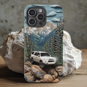 May include: A phone case featuring a white SUV driving over rocks, with a backdrop of mountains, trees, and clouds. The case has a textured, 3D-like appearance, with a detailed landscape design.