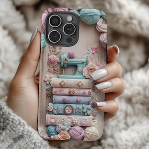 Op de afbeelding: Een telefoonhoesje met een pastelkleurig ontwerp met een naaimachine, boeken, bloemen en bollen wol. De hoes heeft een lichtroze achtergrond met een 3D-effect. De naaimachine is lichtblauw en de boeken zijn versierd met bloemmotieven.