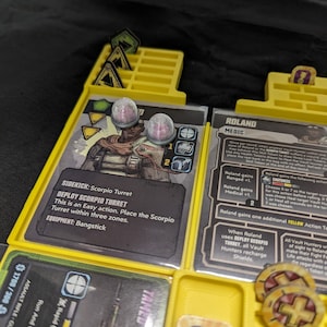 Puede incluir: Un tablero de juego amarillo y negro con una carta de personaje para Roland, un médico. La carta muestra a Roland con una bolsa médica y una jeringa. El texto de la carta dice "Roland gana Ranged +1. Roland gana Medical +1. Roland gana un token de acción AMARILLO adicional. Cuando Roland usa DEPLOY SCORPIO TURRET, todos los cazadores de Vault recargan sus escudos."