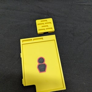 May include: A yellow plastic tray with a black figure and a separate yellow plastic piece with multiple rectangular slots.