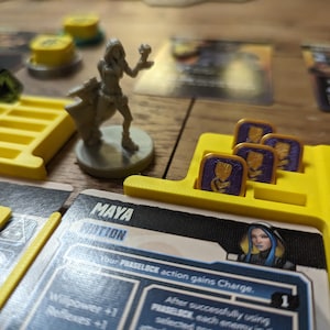 May include: A yellow plastic game board with a card that reads "MAYA MOTION" and the text "Your PHASELOCK action gains Charge." The card features a woman with blue hair and a purple and gold token.