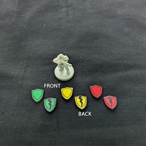 Può includere: Una figura in miniatura di un soldato che tiene un'arma, in piedi su una base rotonda. Ci sono sei piccoli scudi con colori e disegni diversi, tra cui verde, giallo, rosso e nero.