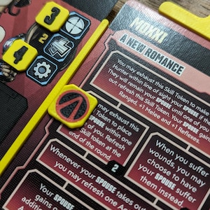 May include: Close-up of a board game component with cards and yellow tokens. The cards have text and are red and white. The yellow tokens have numbers and symbols. The game is set on a wooden surface.