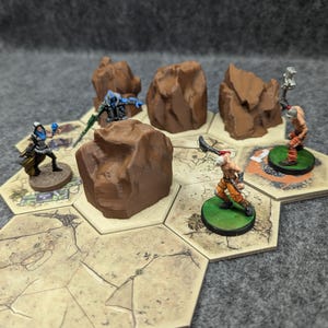 May include: A tabletop game set with four miniature figures and five brown rock terrain pieces. The figures are painted in blue, green, orange, and brown. The figures are standing on hexagonal game tiles.