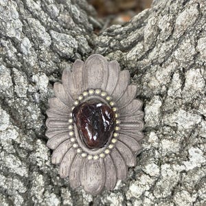May include: A decorative, gray-brown floral-shaped object with a dark, reddish-brown stone center. The stone is surrounded by a ring of small, gold-colored beads. The object is set against a textured, gray tree bark background.