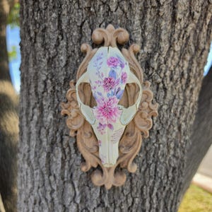 May include: An animal skull featuring a floral pattern in shades of pink and purple, mounted on a light brown ornate oval plaque. The skull displays detailed botanical illustrations of flowers and leaves across its surface.