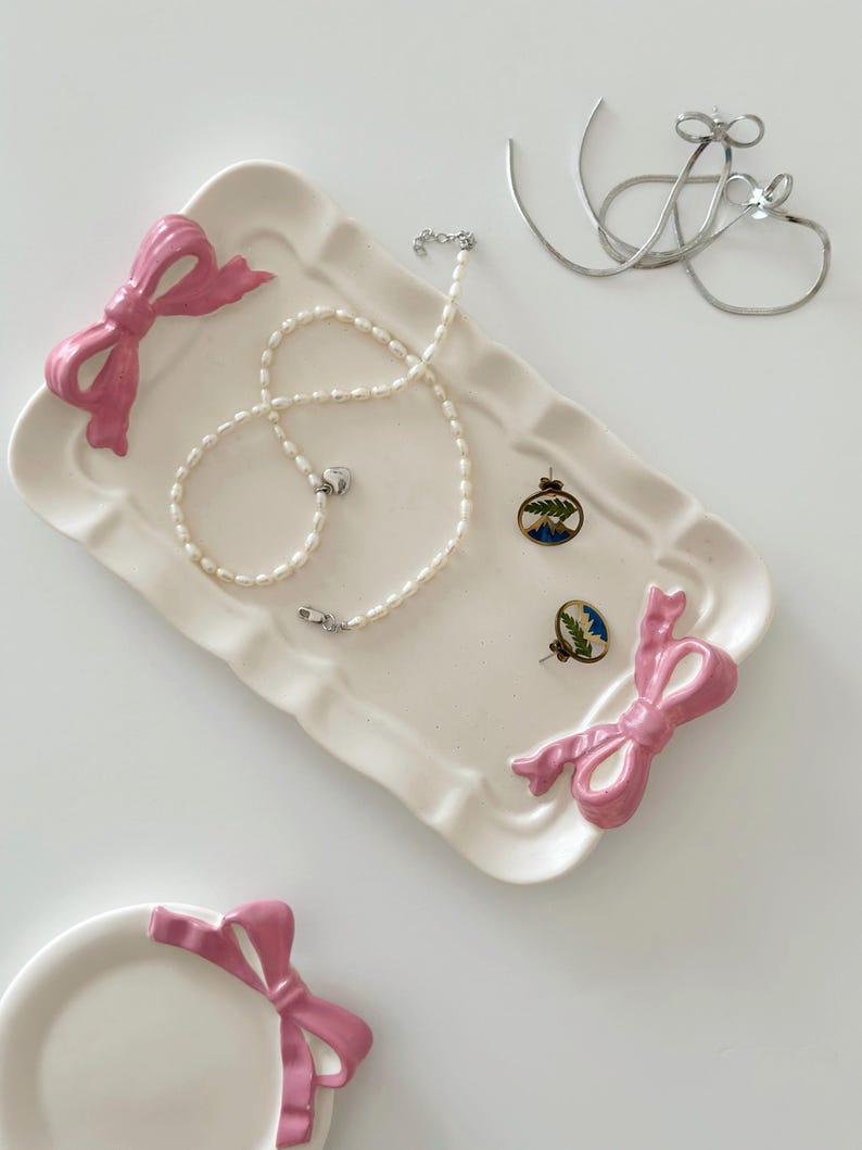 May include: A white ceramic tray with pink ribbon accents. The tray features a pearl necklace, a pair of earrings with a blue and green design, and a silver chain.