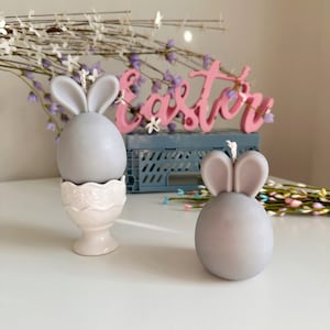Może przedstawiać: Dwie szare świeczki w kształcie królika, jedna w białym stojaku na jajka, z napisem "Easter" w różowych literach w tle.