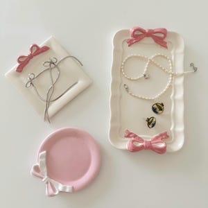 May include: Three pink ceramic dishes with white ribbon accents. The dishes are designed to hold jewelry, with one dish shaped like a square, one shaped like a rectangle, and one shaped like a circle.