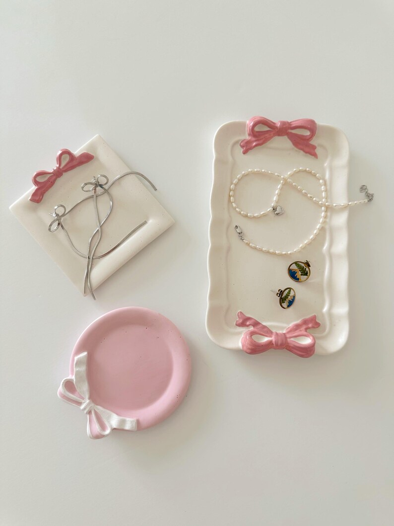 May include: Three pink ceramic dishes with white ribbon accents. The dishes are designed to hold jewelry, with one dish shaped like a square, one shaped like a rectangle, and one shaped like a circle.