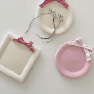 May include: Three ceramic trinket dishes with pink bows. One dish is round and white, one is round and pink, and one is square and white.