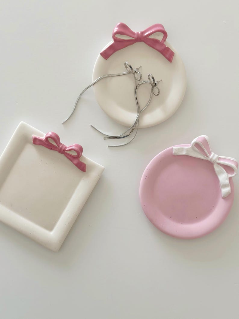 May include: Three ceramic trinket dishes with pink bows. One dish is round and white, one is round and pink, and one is square and white.