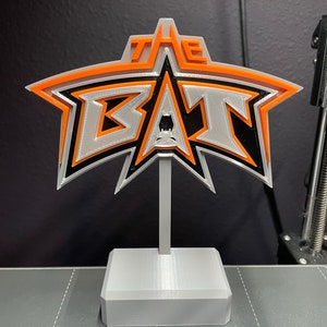 May include: A 3D printed white, black, and orange star-shaped sign with the text "THE BAN" on it. The sign is mounted on a white base.