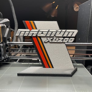 May include: A 3D printed model of a silver "Magniun XL200" logo with red, orange, and black stripes. The logo is mounted on a black base.