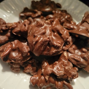 May include: A white plate piled high with chocolate-covered peanut clusters. The clusters are irregular in shape and have a glossy, dark chocolate coating.
