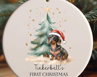 May include: Round ceramic Christmas ornament featuring a dachshund wearing a Santa hat, next to a Christmas tree. The ornament has the text "Tinkerbell's FIRST CHRISTMAS 2025" in black script. Gold stars and a gold star on top of the tree complete the design.