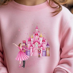 May include: Pink sweatshirt featuring a fairytale design. The graphic includes a pink castle, a ballerina, a prince, and a nutcracker. The design is embellished with stars and sparkles, creating a whimsical aesthetic.