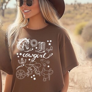 K&ouml;nnte beinhalten: Braunes T-Shirt mit einem wei&szlig;en Grafikdesign aus einem Pferd, Cowboystiefeln, einem Hufeisen, einem Lasso, einem Wagenrad, einem Cowboyhut und einem Sattel. Der Text "cowgirl" ist ebenfalls im Design enthalten.