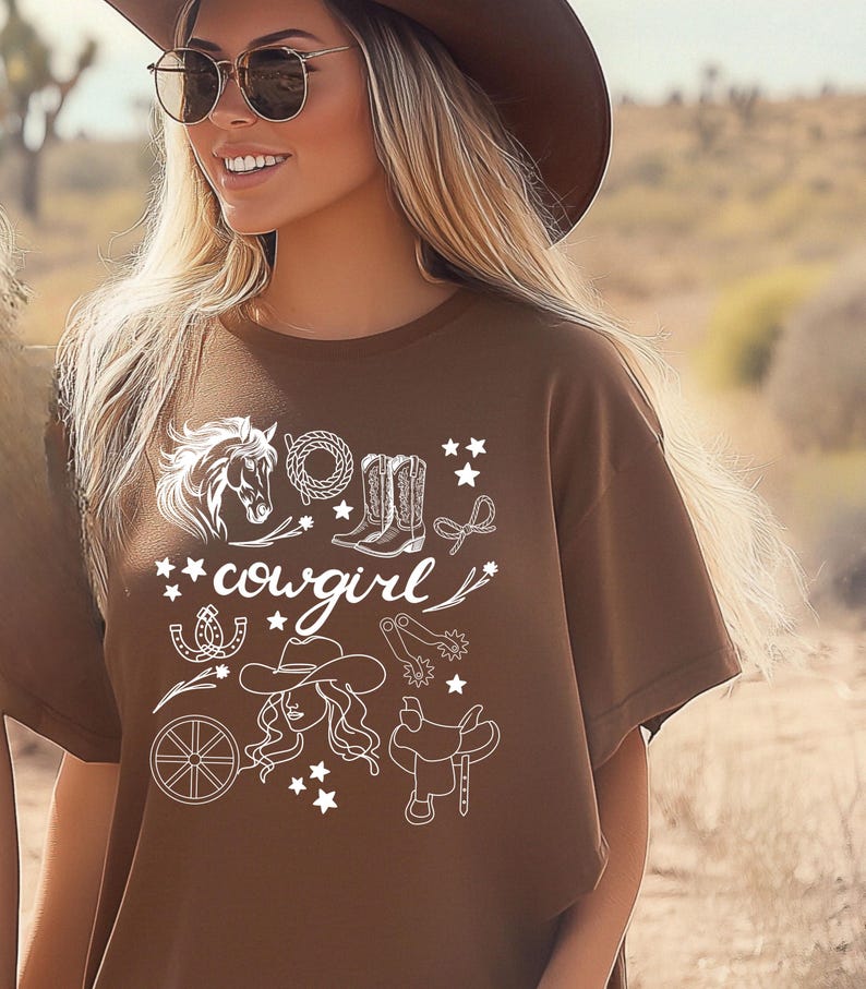 K&ouml;nnte beinhalten: Braunes T-Shirt mit einem wei&szlig;en Grafikdesign aus einem Pferd, Cowboystiefeln, einem Hufeisen, einem Lasso, einem Wagenrad, einem Cowboyhut und einem Sattel. Der Text "cowgirl" ist ebenfalls im Design enthalten.