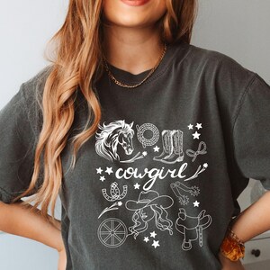 K&ouml;nnte beinhalten: Ein dunkelgraues T-Shirt mit einem wei&szlig;en Grafikdesign einer Cowgirl mit einem Pferd, Stiefeln, einem Hufeisen, einem Sattel und Sternen. Der Text "cowgirl" ist in Wei&szlig; geschrieben.