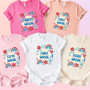 May include: Five colorful t-shirts with grade level designs. Each shirt features a grade level, such as "Fourth Grade" or "First Grade", surrounded by rainbows, flowers, and hearts. The shirts are in shades of pink, white, and beige.