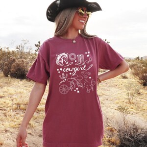 K&ouml;nnte beinhalten: Ein maroonfarbenes T-Shirt mit einem wei&szlig;en Grafikdesign einer Cowgirl mit einem Pferd, Stiefeln und anderen Western-Motiven. Der Text "cowgirl" ist im Design enthalten.