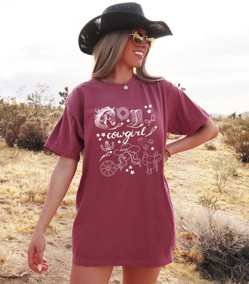 K&ouml;nnte beinhalten: Ein maroonfarbenes T-Shirt mit einem wei&szlig;en Grafikdesign einer Cowgirl mit einem Pferd, Stiefeln und anderen Western-Motiven. Der Text "cowgirl" ist im Design enthalten.