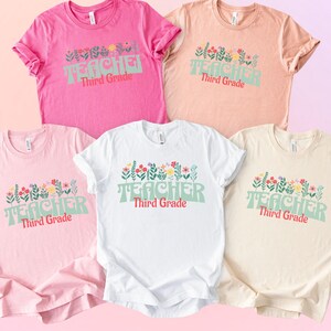 May include: Five t-shirts in various colors: pink, peach, white, light pink, and cream. Each shirt features the word "TEACHER" in green, with "Third Grade" in red, and a floral design.