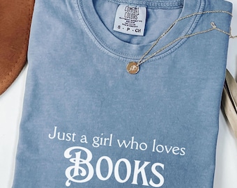 May include: A light blue t-shirt with the text "Just a girl who loves BOOKS" in white. The shirt has a distressed look and is made of a soft, comfortable fabric.