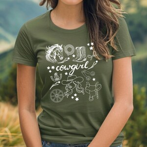 K&ouml;nnte beinhalten: Ein gr&uuml;nes T-Shirt mit einem wei&szlig;en Grafikdesign einer Cowgirl mit einem Pferd, Stiefeln, Sternen und anderen Western-Motiven. Der Text "cowgirl" ist im Design enthalten.