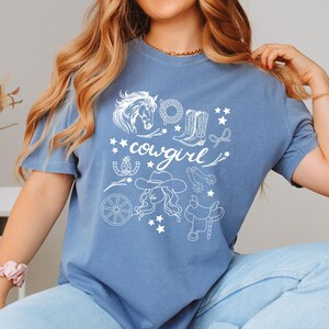 K&ouml;nnte beinhalten: Ein blaues T-Shirt mit einem wei&szlig;en Grafikdesign einer Cowgirl mit einem Pferd, Stiefeln, Sternen und anderen Western-Motiven. Der Text "cowgirl" ist auf dem Shirt zu sehen.