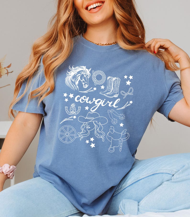 K&ouml;nnte beinhalten: Ein blaues T-Shirt mit einem wei&szlig;en Grafikdesign einer Cowgirl mit einem Pferd, Stiefeln, Sternen und anderen Western-Motiven. Der Text "cowgirl" ist auf dem Shirt zu sehen.