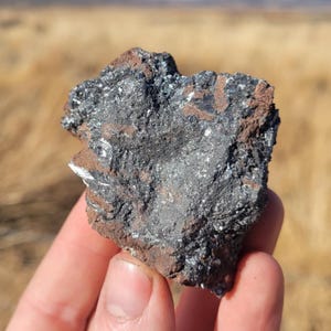 May include: A close-up of a dark gray mineral specimen with a metallic sheen. The rock has a rough, textured surface with areas of reddish-brown and small, reflective crystalline inclusions. The specimen is held in a hand against a blurred outdoor background.