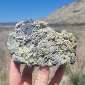 Puede incluir: Primer plano de una roca gris oscura con depósitos minerales verdes y blancos. La roca está siendo sostenida en la mano de una persona.