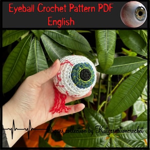 Op de afbeelding: Een gehaakt oog met rode aderen en een groene iris. Het oog wordt in een hand gehouden tegen een groene, bladerige achtergrond. De tekst "Organs collection by Katzcreationcrochet" is zichtbaar onderaan de afbeelding.