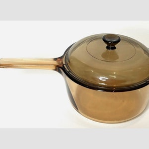 May include: Amber-tinted glass saucepan with a matching lid and a long handle. The lid has a dark knob. The pan is designed for stovetop cooking. The glass material allows for easy monitoring of food while cooking.