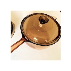 May include: A clear, amber-coloured glass saucepan with a matching lid. The saucepan has a long handle and a round, transparent lid with a small knob. The cookware is designed for stovetop use.