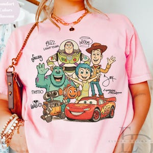 Puede incluir: Una camiseta de color rosa claro con un estampado gr&aacute;fico de personajes de Disney y Pixar, incluidos Buzz Lightyear, Woody, Sulley, Joy, Nemo, Wall-E y Lightning McQueen, con sus nombres escritos en una fuente manuscrita estilizada.