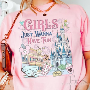May include: Pink t-shirt with the phrase "GIRLS JUST WANNA HAVE FUN" and a colorful graphic featuring a castle, Mickey Mouse, popcorn, and a drink. The shirt has short sleeves and a crew neck.