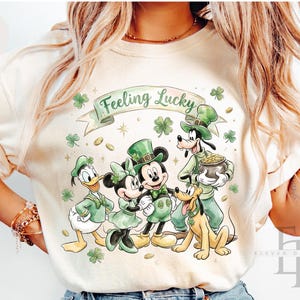 May include: Ivory-colored t-shirt featuring a St. Patrick's Day design with Disney characters. The graphic includes Mickey and Minnie Mouse, Donald Duck, Goofy, and Pluto, all dressed in green attire. The phrase "Feeling Lucky" is displayed above the characters.