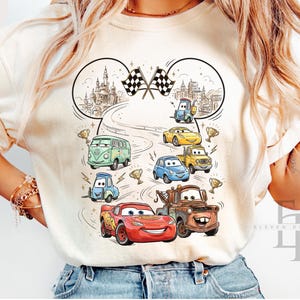 May include: Ivory-colored t-shirt featuring a colorful graphic of cartoon cars racing. The design includes the text "Comfort Colors Ivory" and a cityscape with checkered flags. The cars are in various colors, including red, yellow, and blue.