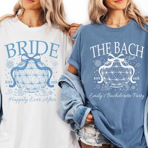 May include: Two t-shirts, one white and one blue, with matching designs. The white shirt says "BRIDE" in blue script above a geometric design. The blue shirt says "THE BACH CLUB" above a similar design. Both shirts have "EST. 2026".