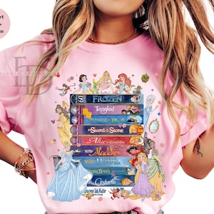 Puede incluir: Camiseta rosa claro con una pila de t&iacute;tulos de pel&iacute;culas animadas cl&aacute;sicas, incluyendo "Frozen" y "Tangled." El dise&ntilde;o incorpora personajes de princesas Disney y el texto "Comfort Colors Blossom."