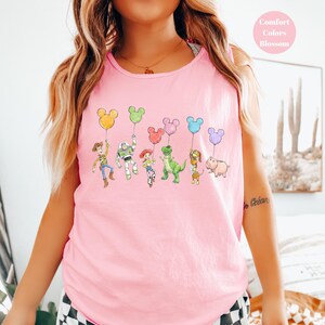May include: Pink tank top featuring a colorful graphic of cartoon characters holding Mickey Mouse-shaped balloons. The characters include Woody, Buzz Lightyear, Jessie, Rex, Slinky Dog, and Hamm. The text "Comfort Colors Blossom" is in a pink circle.
