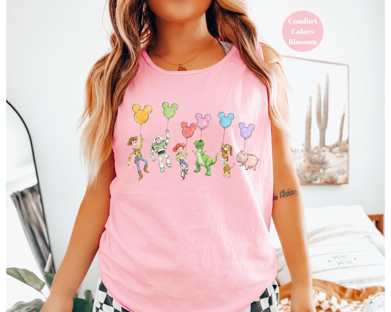 May include: Pink tank top featuring a colorful graphic of cartoon characters holding Mickey Mouse-shaped balloons. The characters include Woody, Buzz Lightyear, Jessie, Rex, Slinky Dog, and Hamm. The text "Comfort Colors Blossom" is in a pink circle.
