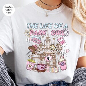 May include: White t-shirt with the text "THE LIFE OF A PARK GIRL" in blue and pink. The shirt features illustrations of a castle, popcorn, a coffee cup, a wand, and other park-themed graphics. The words "Comfort Colors White" are in a circle in the upper left.
