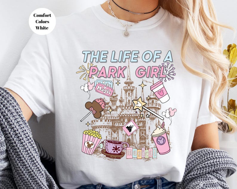 May include: White t-shirt with the text "THE LIFE OF A PARK GIRL" in blue and pink. The shirt features illustrations of a castle, popcorn, a coffee cup, a wand, and other park-themed graphics. The words "Comfort Colors White" are in a circle in the upper left.