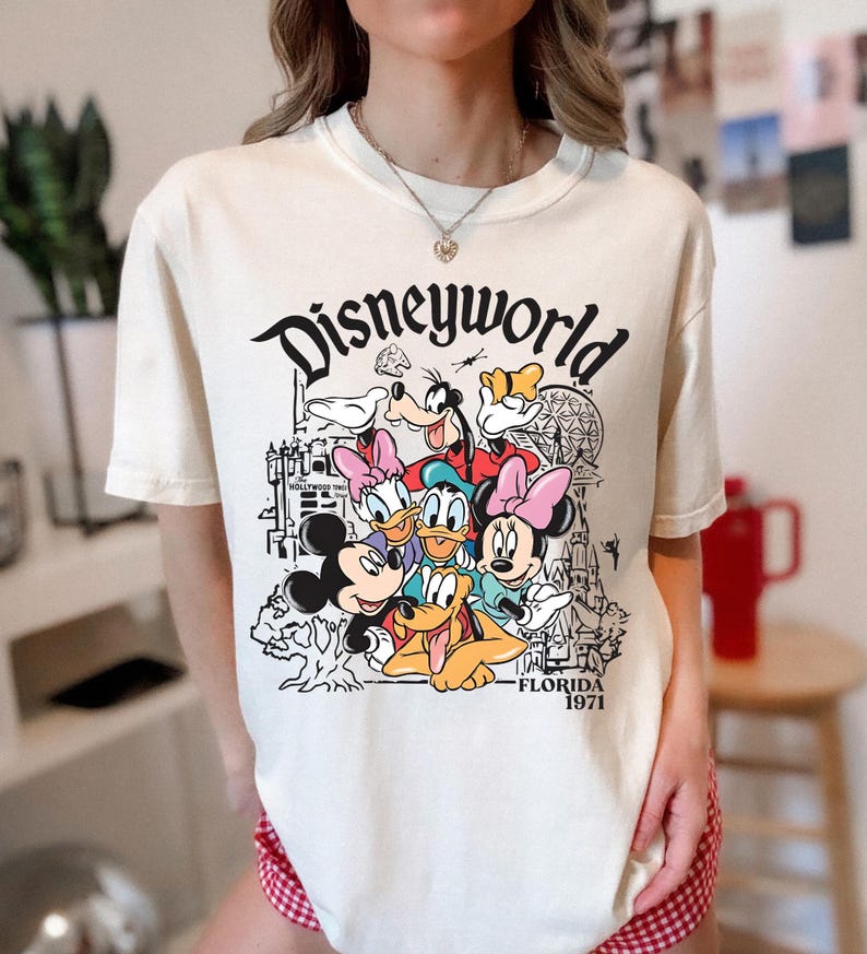 May include: Cream-coloured t-shirt with a graphic of Disney characters such as Mickey Mouse, Minnie Mouse, Donald Duck, and Goofy. The word "Disneyworld" is above the characters, with "Florida 1971" below.